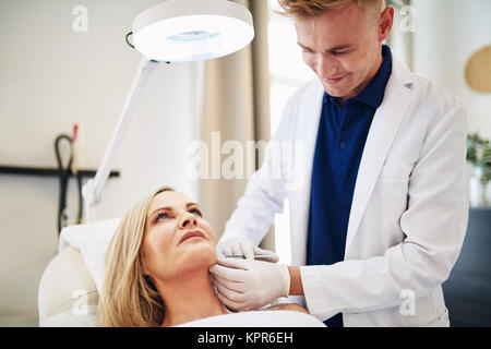 Young male doctor focused on performing a injection to the chin of a mature woman lying on a table in a beauty clinic Stock Photo