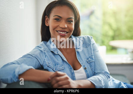 Casually dressed young African woman smiling while sitting alone on the sofa in her living room at home Stock Photo