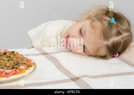 Little girl with protruding tongue rested her head on the table and looks at the pizza Stock Photo