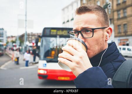 Morning coffee on the city street. Young man drinking hot coffee from the disposable cup and listening music in earphones. Stock Photo