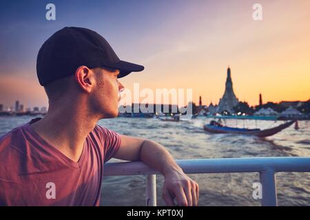 Young man (tourist) on the boat watching the traditional boats against temple Wat Arun. Bangkok at the sunset, Thailand. Stock Photo