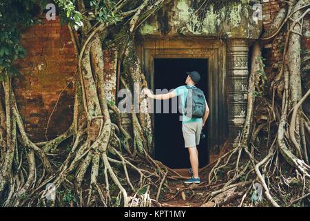 Young man with backpack coming to ancient monuments under the giant roots of the tree near Siem Reap (Angkor Wat) in Cambodia Stock Photo