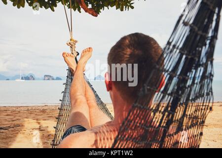Young man (tourist) relaxing in the hammock on the sand beach - selective focus on foot. Koh Yao Noi island situated between Phuket and Krabi in Thail Stock Photo