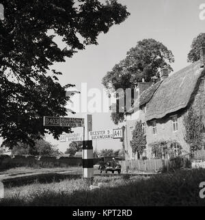 1950s, historical, England, road sign showing the B403 to Oxford or the A41 to Bicester as a motorcar of this era passes a thatched cottage. Stock Photo