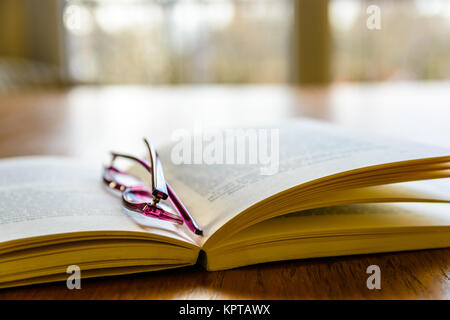 Close-up view of an open pocket book with a pair of reading glasses on it, laying flat on a wooden table in front of a window. Stock Photo