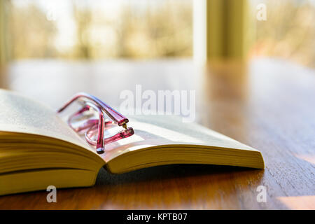 Close-up view of an open pocket book with a pair of reading glasses on it, laying flat on a wooden table in front of a window. Stock Photo