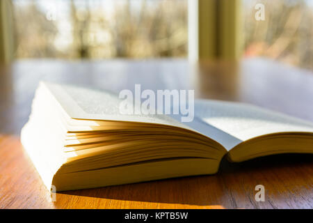 Close-up view of an open pocket book laying flat on a wooden table and illuminated by a ray of sunlight, in front of a window. Stock Photo