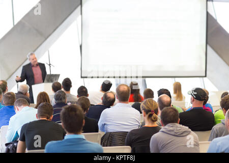 Man giving presentation in lecture hall. Male speeker having talk at public event. Participants listening to lecture. Rear view, focus on people in audience. Stock Photo