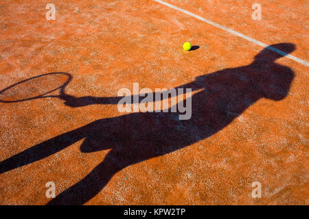 Shadow of a tennis player in action on a tennis court (conceptual image with a tennis ball lying on the court and the shadow of the player positioned in a way he seems to be playing it) Stock Photo