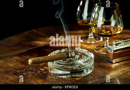 Glass ashtray with smoking cigar by two glasses filled with bourbon besides fancy lighter on a wood table Stock Photo
