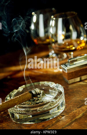 Tilted angle view of cigar sitting in glass ashtray by fancy lighter and two goblets filled with bourbon Stock Photo