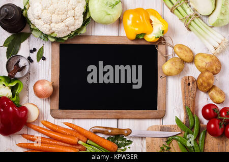 Blank rectangular chalkboard with wooden frame surrounded by fresh vegetables as onion, carrots, pepper, carrots, tomatoes, cauliflower, herbs and spices on a rustic table, close up from above Stock Photo