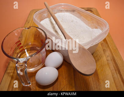 Baking utensils, a glass measuring cup, a container of flour with a wooden spoon and two eggs on a wooden cutting board sitting on an orange counter t Stock Photo