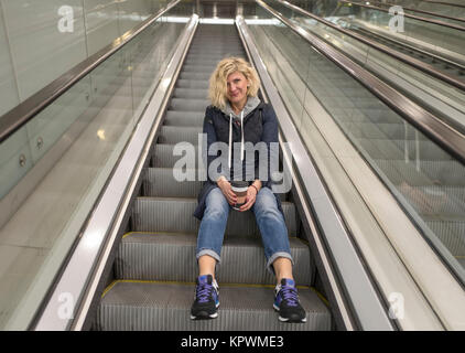 Blonde girl is seating on the stairs of escalator Stock Photo