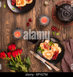 Valentine's day flat lay with delicious pancakes in form of heart, green tea, black teapot, candles and roses. Valentines day concept greeting card. T Stock Photo