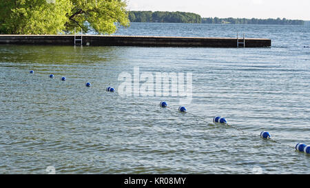 Buoys strung together by rope along beautiful blue lake to create safe swimming area for swimmers.  Dock with ladder in background.  No people. Stock Photo