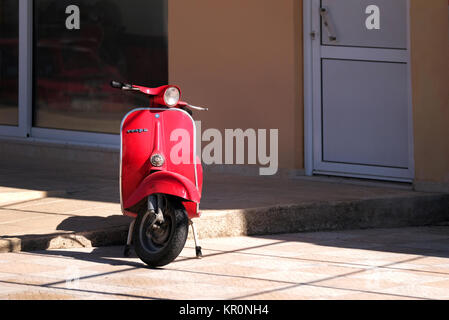 An old red Vespa scooter parked on a stand on the street. Despite its age the scooter is in good condition and bright sunlight shows off its red paint Stock Photo