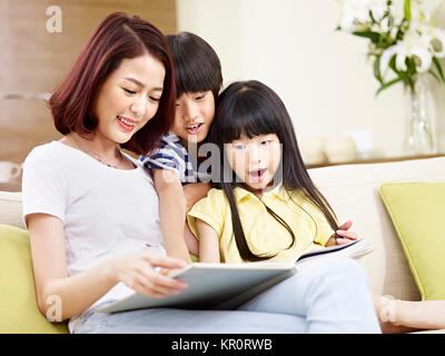 young asian mother sitting on couch at home reading story to two little children. Stock Photo