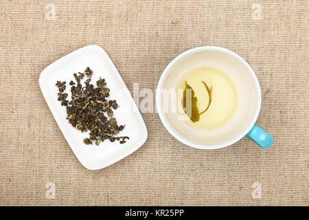 Big empty cup of green tea with leaf on bottom and white plate with dried green tea leaves on flax canvas tablecloth, close up, elevated top view, dir Stock Photo