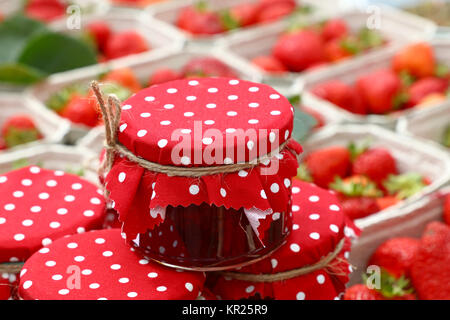 Close up of small glass jars of berry jam with red dotted rustic top lid textile decoration and twine bow over background of fresh strawberry on retai Stock Photo