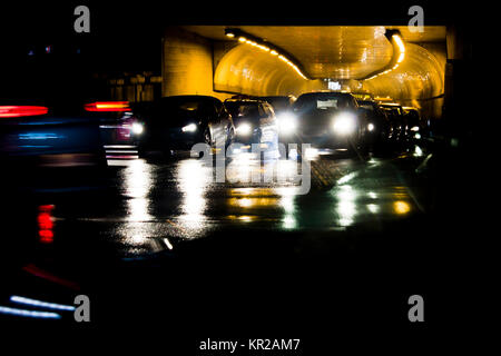 Night traffic on rainy city streets. Cars queued at tunnel exit waiting at intersection while driving vehicles moving past, front view with display da Stock Photo