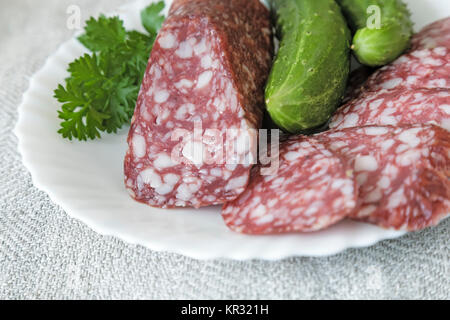 On the tablecloth of the table is a plate of sliced sausages, cucumbers and parsley. Stock Photo