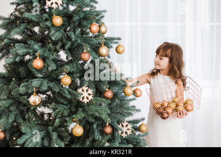 Beautiful little girl decorates huge green Christmas tree for holiday celebration. Horizontal color photography. Stock Photo