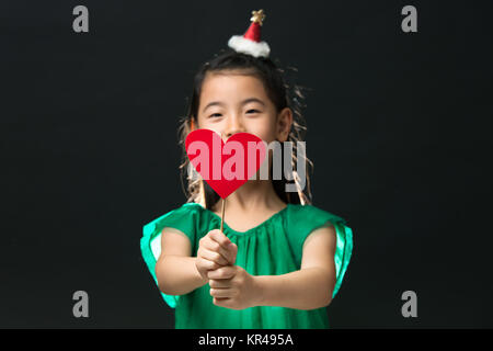 cute asian girl child dressed in a green dress holding a Christmas ornament and a heart stick on a black background. Face-masked Stock Photo