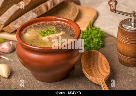 fresh transparent fish soup with sturgeon, potatoes in clay pot, decorated garlic, parsley leaves, cutted rye bread, wooden spoon and grinder, ethnic  Stock Photo