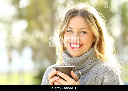 Happy woman wearing a grey jersey looking at camera holding a cup of coffee Stock Photo