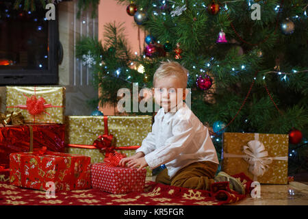 Cute blonde hair little boy near the fireplace and gifts under Christmas tree. Stock Photo