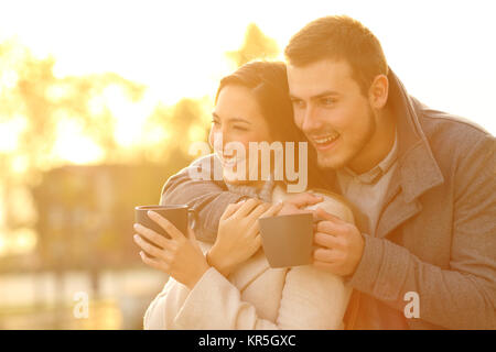 Portrait of a happy couple looking away holding coffee mugs in winter at sunset Stock Photo