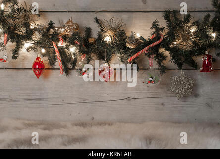 Christmas pine tree garland with red baubles, candy canes and ornaments, hanging on old wooden board background, standing on sheepskin Stock Photo