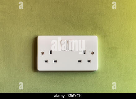 UK domestic 3 pin double plug electrical socket in white plastic on a green coloured wall Stock Photo