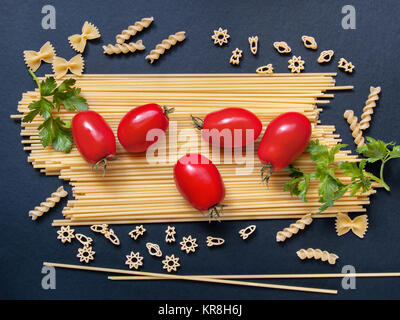 Spaghetti with tomatoes, greens and other figured pasta in the form of bows, spirals, figures of rockets and stars for cooking on a dark background Stock Photo