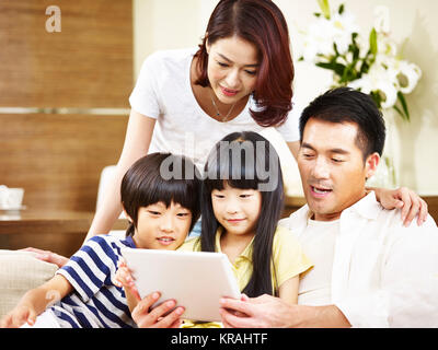 asian parents mother and father and two children son and daughter sitting on family couch using digital tablet together. Stock Photo