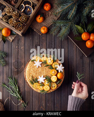 Woman decorates Christmas Honey cake with rosemary and oranges on brown wooden background. Stock Photo