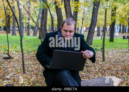 Businessman having a laptop in the park in the fall. Young man sitting on tree in park with laptop on his lap and checks e-mail. A man works and looks Stock Photo