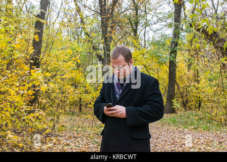 Smiling young man reading sms on phone. A man checks the mail on the phone. Passive autumn a lot of yellow leaves Stock Photo