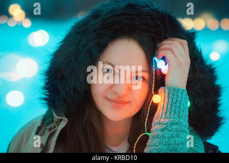 Portrait of a smiling young brunette woman dressed in a winter jacket with a fur hood, holding multicolor string lights near her face. Stock Photo