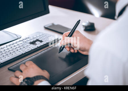 Graphic designer's workspace. Hand on pen tablet. Young smart man in the office. Free black monitor copy space for design. Warm glare of sun light. Stock Photo
