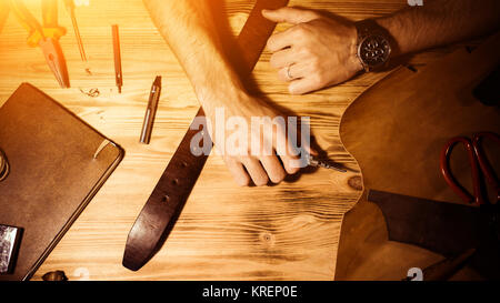 Working process of the leather belt in the leather workshop. Man holding hands on wooden table. Crafting tools on background. Tanner in old tannery. W Stock Photo
