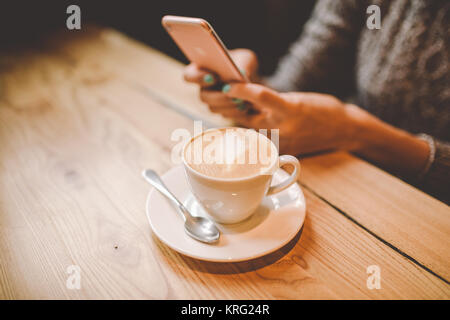 hands close-up of a beautiful young girl uses,types text on a mobile phone at a wooden table near a window and drinks coffee in a cafe decorated with Christmas decor. Dressed in a gray knitted sweater Stock Photo