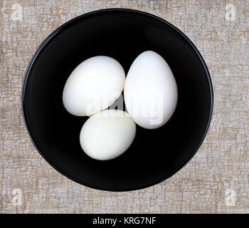 Three large white duck eggs in a black bowl on a textured surface shot from ablove. Stock Photo