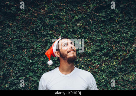 Young brunette man, wearing a christmas cap, over a texture vegetal background, looking up and smiling Stock Photo