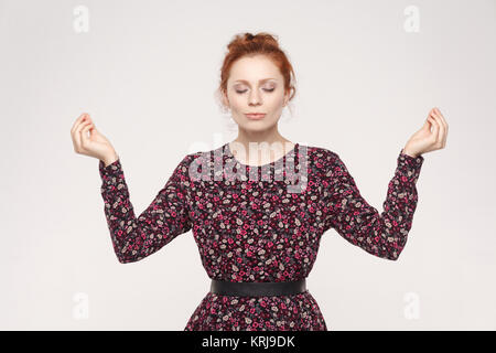 Meditation, religion and spiritual practises. Beautiful  ginger woman doing yoga  indoors at grey wall, keeping eyes closed, holding fingers in mudra  Stock Photo