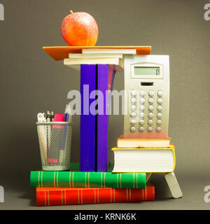 Concept of education. Stack of school books with calculator, marker and pencils and a red apple on top in front of dark gray background Stock Photo