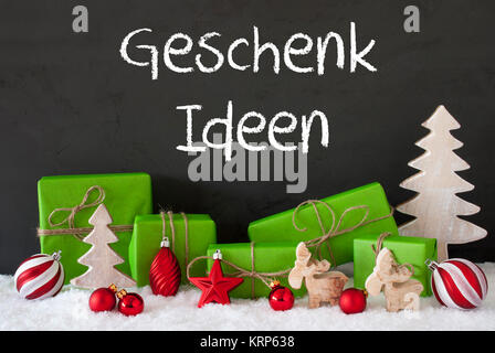 German Text Geschenk Ideen Means Gift Ideas. Green Gifts Or Presents With Christmas Decoration Like Tree, Moose Or Red Christmas Tree Ball. Black Cement Wall As Background With Snow. Stock Photo