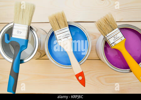 Ready to paint a house. Three tin cans of household paint in a row with brushes on top, light uncolored wooden background. Close up. Top view. Space f Stock Photo