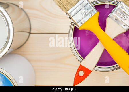 Ready to paint an apartment. A tin can of fuchsia oil paint with crossed brushes on top in close up, light uncolored wooden background. Top view. Spac Stock Photo
